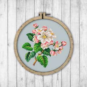 May include: Cross-stitch embroidery of a flowering branch with white and pink blossoms, green leaves, and a brown stem. The embroidery is set in a round wooden hoop against a light blue background. The hoop is displayed on a white wooden surface.