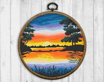 May include: A circular wooden frame containing a colourful cross-stitch design depicting a sunset over a lake with two trees in the foreground. The sky is a gradient of blue, purple and orange, and the lake is a reflective blue with orange and yellow highlights.