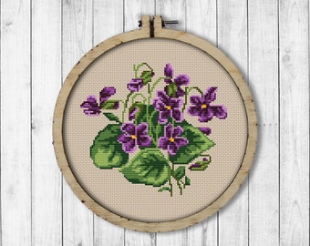 Puede incluir: Un aro de bordado de punto de cruz con un diseño de violetas moradas y verdes. El aro es de madera y tiene un fondo de tela beige.