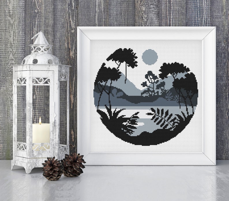 May include: Black and white cross stitch pattern of a circular landscape with trees, mountains, and a lake. The design is framed in a white frame.
