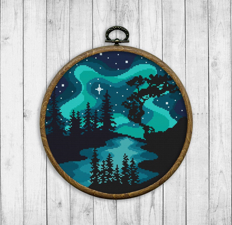 Op de afbeelding: Een ronde houten borduurring met een kruissteekontwerp van een boslandschap onder een blauwe en groene aurora borealis. Het ontwerp bevat een meer, bomen en sterren.
