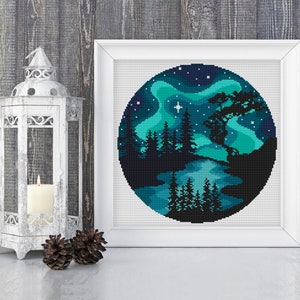 Op de afbeelding: Een rond kruissteekpatroon dat een donkerblauwe nachthemel met een groene aurora borealis, een meer en een silhouet van een bos met bomen afbeeldt. Een enkele witte ster schijnt aan de hemel.