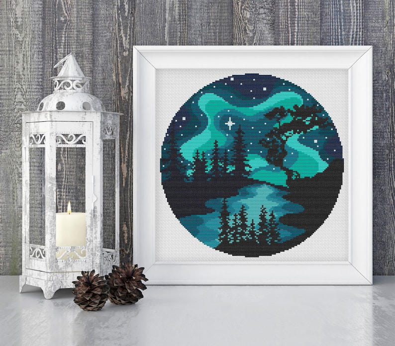 Op de afbeelding: Een rond kruissteekpatroon dat een donkerblauwe nachthemel met een groene aurora borealis, een meer en een silhouet van een bos met bomen afbeeldt. Een enkele witte ster schijnt aan de hemel.