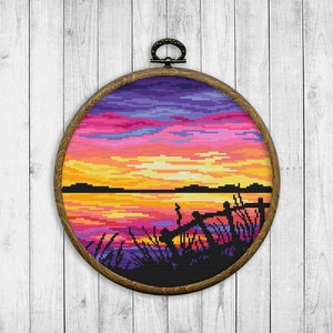 Peut inclure: Une broderie au point de croix encadrée représentant un coucher de soleil sur un lac avec une silhouette de quai et de roseaux au premier plan. Le ciel est un mélange vibrant de rose, de violet, d'orange et de jaune.