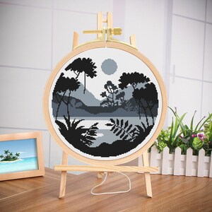 May include: A black and white cross-stitch embroidery of a forest scene with a lake and a moon in the sky. The embroidery is mounted in a wooden hoop and displayed on a wooden easel.