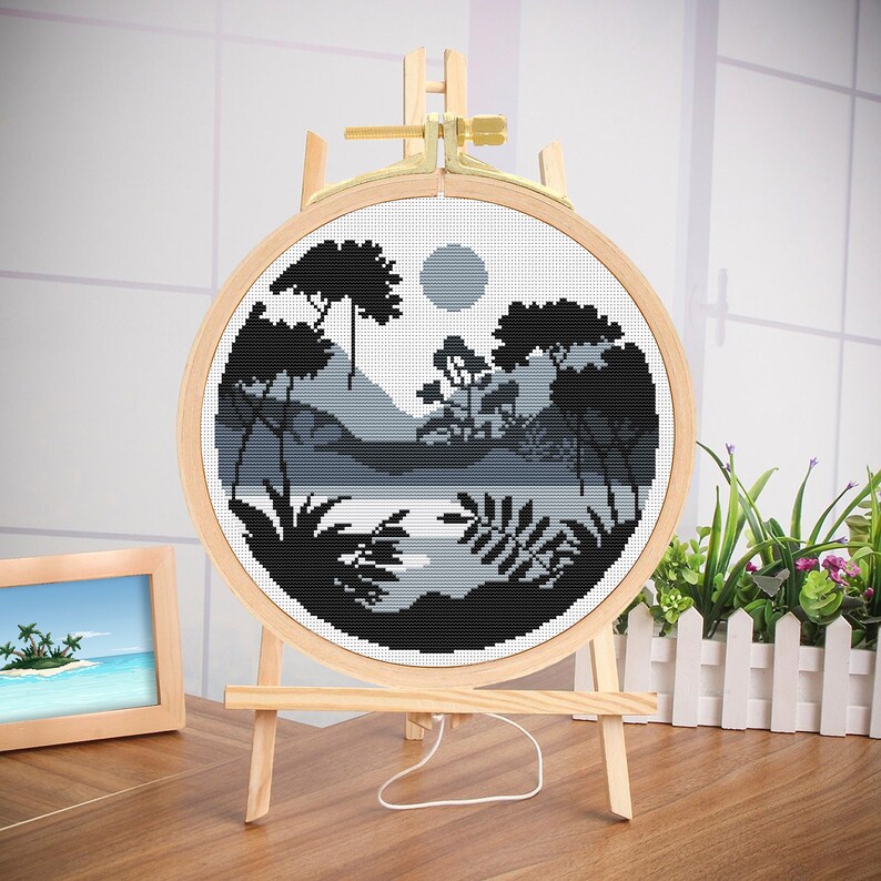 May include: A black and white cross-stitch embroidery of a forest scene with a lake and a moon in the sky. The embroidery is mounted in a wooden hoop and displayed on a wooden easel.