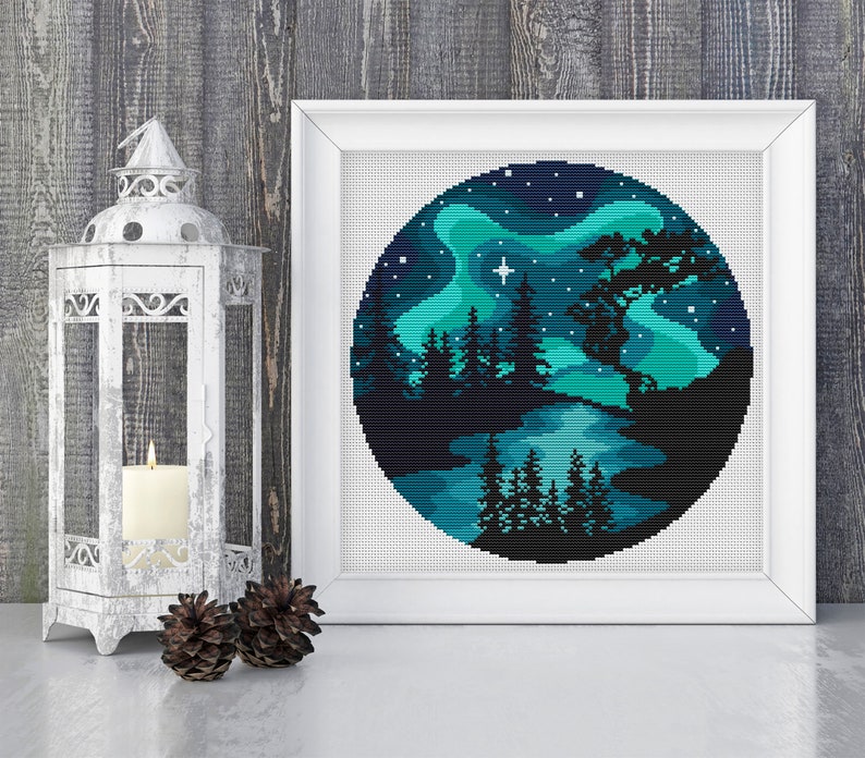 May include: A round cross-stitch pattern depicting a dark blue night sky with a green aurora borealis, a lake, and a silhouette of a forest with trees. A single white star shines in the sky.