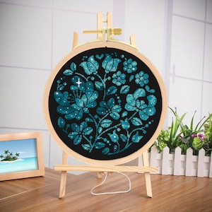 May include: A round embroidery hoop with a blue and black floral design. The design features a cluster of flowers with a star in the center. The hoop is on a wooden stand.