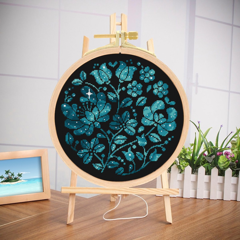 May include: A round embroidery hoop with a blue and black floral design. The design features a cluster of flowers with a star in the center. The hoop is on a wooden stand.