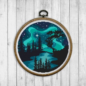 Op de afbeelding: Een borduurring met kruissteek met een ontwerp van een boslandschap onder een sterrenhemel met een groen-blauwe aurora borealis. De bomen zijn gesilhouetteerd tegen de hemel.