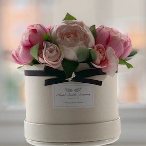 May include: A white round box with a black ribbon and a label that reads "Angel Candle Company Luxury Handmade Peony & Blush Suede" is filled with pink and white artificial flowers.