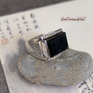 May include: A silver ring with a rectangular black stone center and small clear stones along the top edge. The ring is set on a gray stone, with the text "GoodFortuneWithZ" visible in the background.