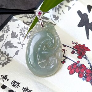 May include: A jade pendant with a swirling design and a silver bail. The pendant is a light green color and has a small, heart-shaped ruby accent. The pendant is displayed on a patterned fabric.