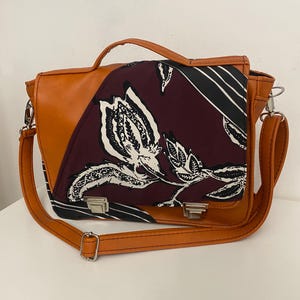 May include: A brown leather messenger bag with a floral patterned flap. The bag has a top handle and a shoulder strap. The flap features a burgundy, black, and white floral design. Silver-coloured clasps secure the bag.