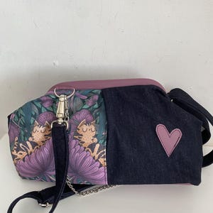 May include: A crossbody bag featuring a purple top panel and a dark blue denim side panel with a purple heart patch. The other side displays a pattern of sleeping cats amidst purple flowers and green foliage, complete with a dark blue strap and silver hardware.