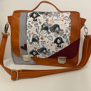 May include: A brown satchel-style shoulder bag with a flap closure. The flap features a white panel with a repeating illustration of women, trees, and umbrellas. The bag has a handle and a detachable shoulder strap. A black and white striped panel is on the left side.