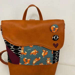 May include: A tan backpack with a curved flap featuring three decorative buttons: one with a fox, one with a black and white geometric pattern, and one purple heart. The body includes panels of teal fabric with orange fox prints and black diamond patterns.