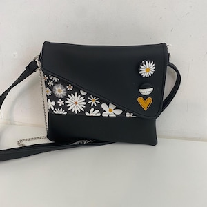 Può includere: Borsa a tracolla nera con un accento a stampa di margherite e bottoni decorativi. La borsa ha una tracolla a catena argentata e una chiusura con patta. Il design include un bottone a margherita bianca e un bottone a cuore giallo.