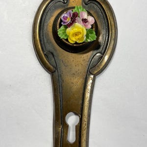 May include: A vintage brass keyhole cover with a floral design and a decorative butterfly. The floral design features a yellow rose with pink and purple flowers. The butterfly is made of gold metal and has a filigree design.