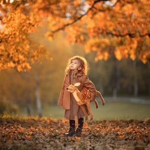 May include: A young girl wearing a brown coat and dress holds a plush fawn toy. She is standing in a field of fallen autumn leaves, looking up at the sky.