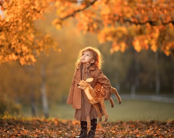 May include: A young girl in a brown coat and dress holds a plush fawn toy. She is standing in a field of fallen autumn leaves, looking up at the sky.