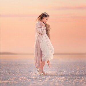 Puede incluir: Una persona con un abrigo rosa claro y un vestido blanco, de pie sobre una superficie texturizada. La persona lleva sandalias doradas. El fondo presenta un cielo de colores pastel suaves.