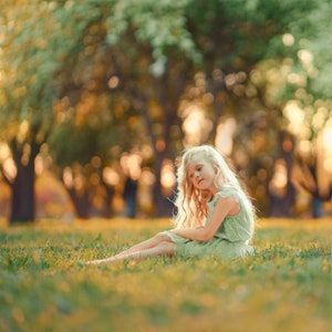 May include: A young girl with long blonde hair sits in a field of green grass. She is wearing a light green dress and has a thoughtful expression on her face.