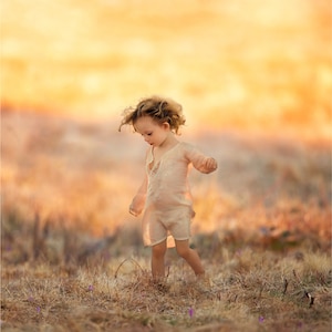 Puede incluir: Un niño pequeño con el pelo rubio rizado camina por un campo de hierba seca. El niño lleva un vestido suelto de color marrón claro. El fondo es una puesta de sol borrosa.