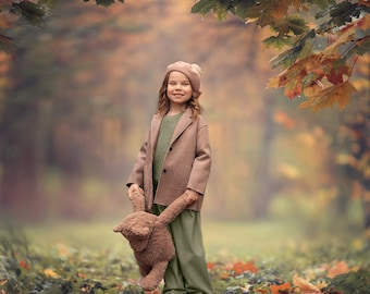 May include: A young girl wearing a brown coat and a pink hat stands in a forest with autumn leaves. She is holding a brown teddy bear.