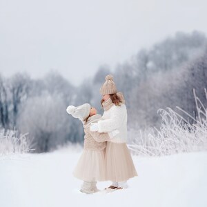 Pode incluir: Duas meninas pequenas com chapéus e casacos de inverno estão em pé em um campo nevado. Elas se olham e sorriem. O fundo é uma floresta desfocada.