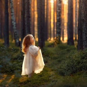 May include: A young girl in a white dress stands in a forest, looking up at the sun shining through the trees.