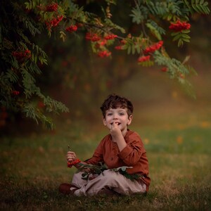Puede incluir: Un niño pequeño está sentado en la hierba bajo un árbol con bayas rojas, sonriendo y comiendo una baya. Lleva una camisa marrón y pantalones beige.