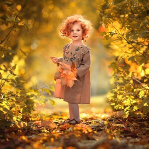 May include: A young girl with red hair and a brown dress stands in a forest with autumn leaves. She is holding a bouquet of yellow leaves.