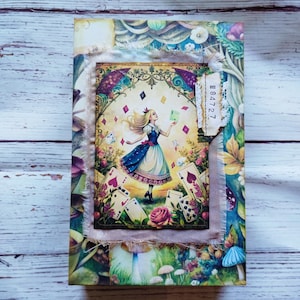 Könnte beinhalten: Ein rechteckiger Bucheinband mit einer zentralen Illustration einer blonden Figur in einem blauen Kleid, umgeben von schwebenden Spielkarten und Blumen. Ein ausgefranstes Stoffstück mit dem aufgedruckten Text No 84727 ist an der rechten Seite befestigt.