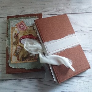 K&ouml;nnte beinhalten: Zwei handgefertigte Journale mit braunen Einb&auml;nden. Ein Journal zeigt ein dekoratives Bild eines Pilzes und eine Vintage-Briefmarke. Das andere Journal ist mit einem wei&szlig;en Band umwickelt. Beide Journale haben eine rustikale, Vintage-&Auml;sthetik.