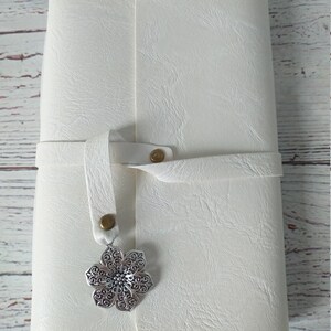 May include: Ivory-colored journal with a textured cover and a strap closure. A silver-colored, ornate flower charm hangs from the strap. The journal is closed and appears to be made of leather or a similar material.