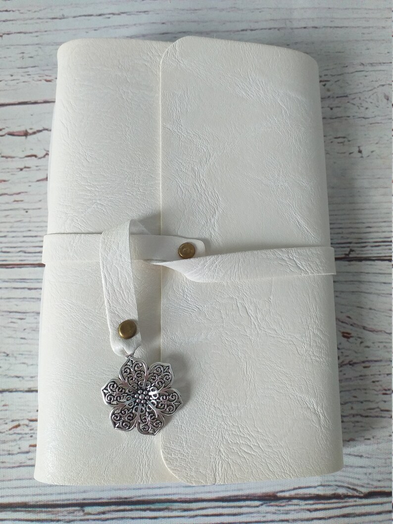 May include: Ivory-colored journal with a textured cover and a strap closure. A silver-colored, ornate flower charm hangs from the strap. The journal is closed and appears to be made of leather or a similar material.