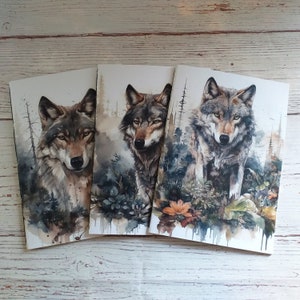 May include: Three watercolor prints featuring detailed portraits of wolves. Each wolf is depicted with realistic fur and piercing eyes, set against a backdrop of a forest with muted greens, browns, and blues. The prints are displayed on a light wood surface.