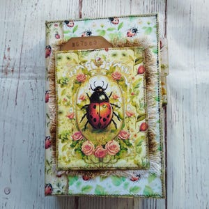 Könnte beinhalten: Ein handgefertigtes Notizbuch mit einem Marienkäfer-Design. Das Cover zeigt ein zentrales Bild eines Marienkäfers in einem floralen Rahmen, umgeben von Rosen und einem Marienkäfermuster. Ein kleines Etikett mit der Nummer "57563" ist angebracht.