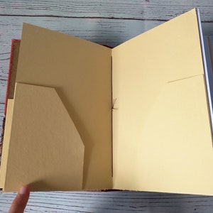 以下が含まれることがあります： ベージュ色のページとポケットが付いた開いたジャーナル。ジャーナルの表紙は赤色で模様が入っています。ページは紐でまとめられています。ジャーナルは開かれており、内側のページとポケットが見えています。