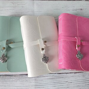 May include: Three leather journals in pastel colors: mint green, cream, and pink. Each journal has a strap closure with a metal heart charm. The journals are closed, with visible stitching on the spine.