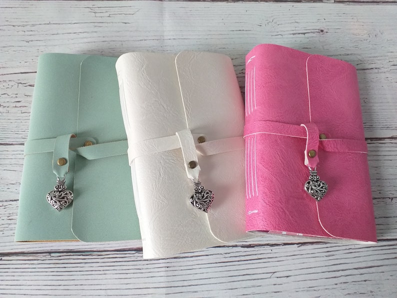 May include: Three leather journals in pastel colors: mint green, cream, and pink. Each journal has a strap closure with a metal heart charm. The journals are closed, with visible stitching on the spine.