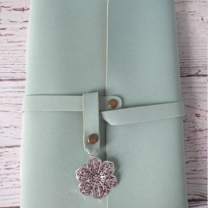 May include: A light teal journal with a wrap-around closure and a decorative silver flower charm. The journal is closed with a leather strap and two brass-colored rivets. The cover has a smooth texture.