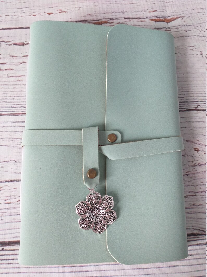 May include: A light teal journal with a wrap-around closure and a decorative silver flower charm. The journal is closed with a leather strap and two brass-colored rivets. The cover has a smooth texture.