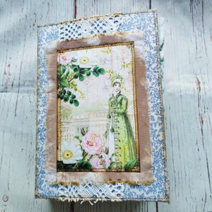 Könnte beinhalten: Ein Notizbuch im Vintage-Stil mit einem floralen Design und einer Frau in einem grünen Kleid auf dem Umschlag. Der Umschlag ist mit Spitze und Stoffresten verziert.