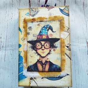 May include: A vintage-style notebook cover with a whimsical illustration of a young wizard wearing a pointed hat and round glasses. The cover is decorated with a distressed, yellow fabric frame and a blue and white patterned background.