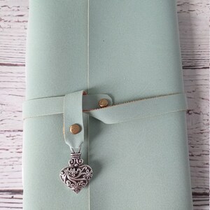 May include: A light green journal with a leather strap closure. The journal has a decorative silver heart charm hanging from the strap. The journal is closed and appears to be new.
