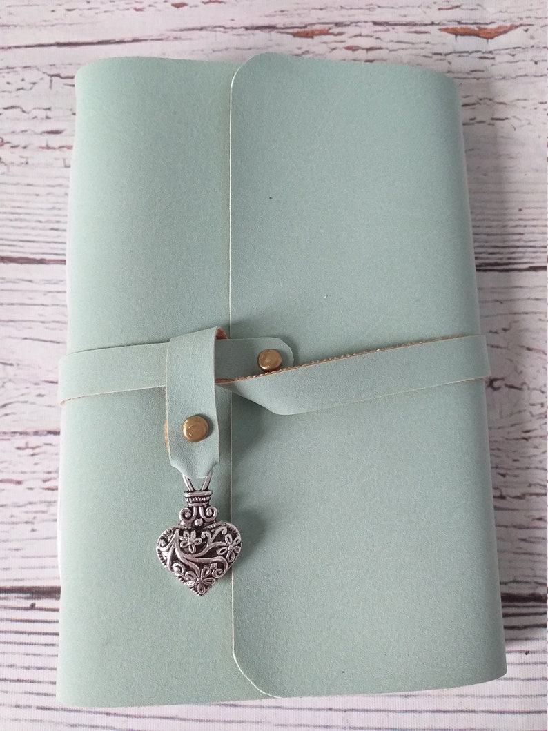 May include: A light green journal with a leather strap closure. The journal has a decorative silver heart charm hanging from the strap. The journal is closed and appears to be new.