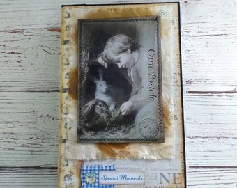 Puede incluir: Un diario hecho a mano con una estética vintage. La portada presenta una imagen en blanco y negro de un niño con conejos, etiquetada como "Carte Postale". Acentuado con tela, una cinta de cuadros azul y las palabras "Special Moments."