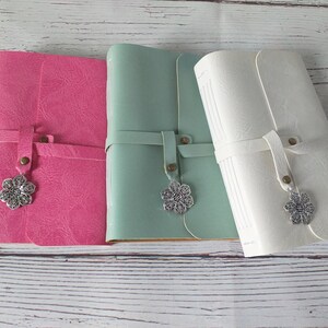 May include: Three leather journals in pink, mint green, and white. Each journal has a strap closure with a decorative silver flower charm. The journals are closed and appear to be new.
